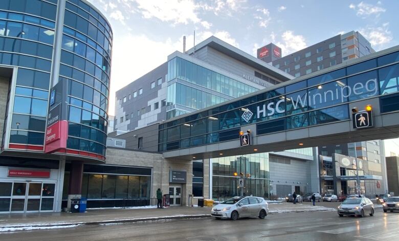 Cars drive down a road past a building complex and under a glass-enclosed pedestrian bridge that says 