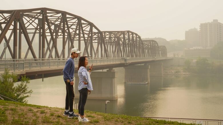 Two people standing by the river in Saskatoon looking at the smoke 