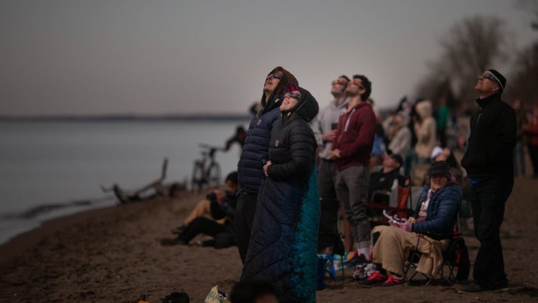 People wearing eclipse glasses look up to the dim sky from the shore of a lake.