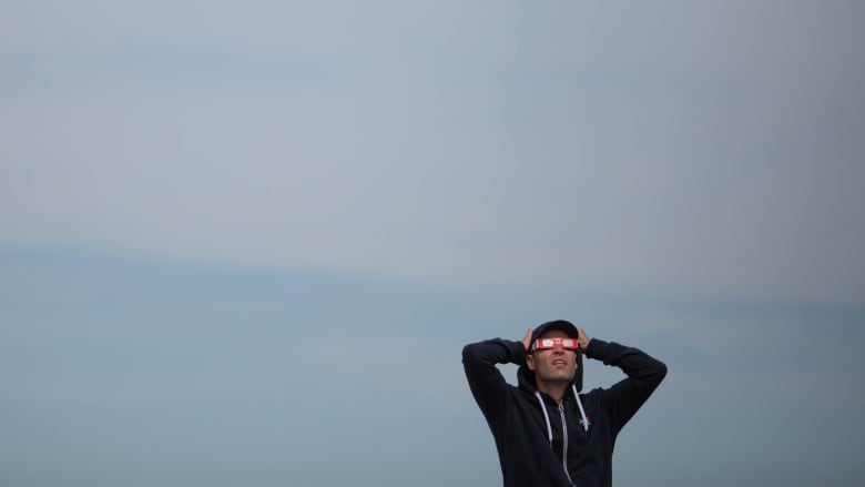 A man in a blue zip-up sweatshirt puts both hands on his head as he watches a solar eclipse through eclipse glasses.