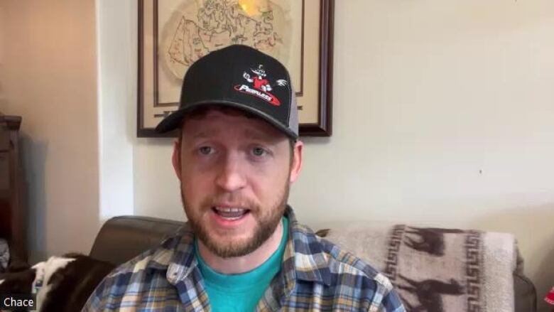 A white man wearing a black hat speaks in front of a sofa.