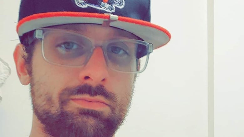 a white male with a  beard wearing glasses, a baseball hat and an orange shirt in close up shot. 