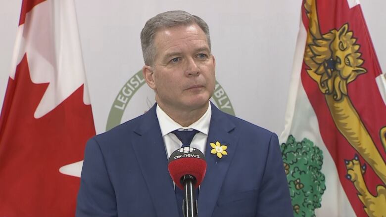 A man in a blue suit and dark tie stands in front of a microphone taking questions from reporters.