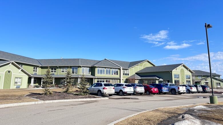 A photo of Diamond House. It has green walls and a grey roof with many cars parked in front of it.