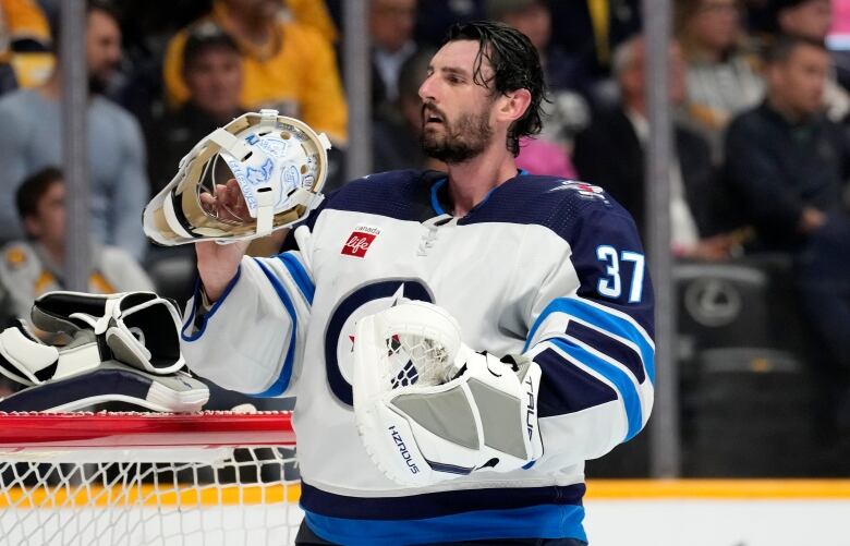 A hockey goalie holds his mask and looks at it