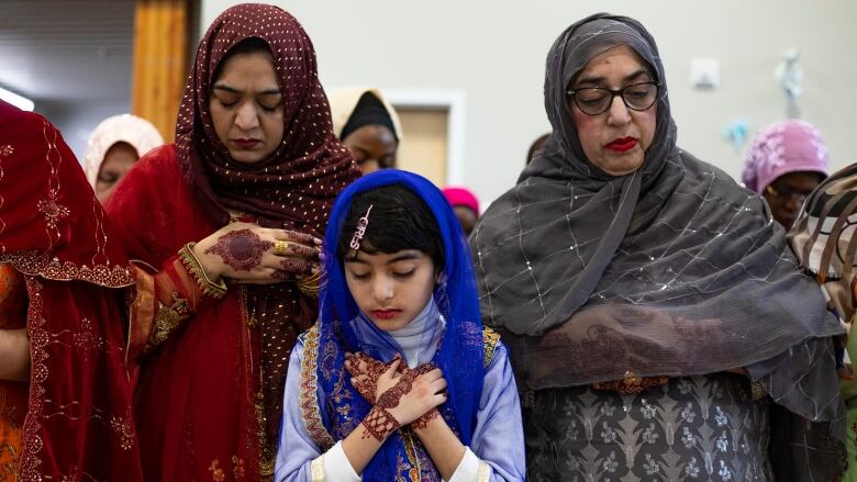 Two women in colourful clothing and head scarves, stand beside each other. A young girl dressed similarly stands in front of them, her arms crossed on her chest.