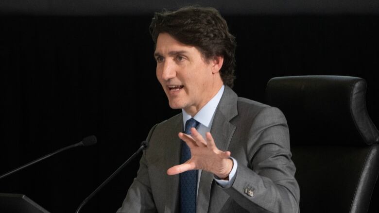 A man in a grey suit gestures with his hand as he speaks into a microphone.