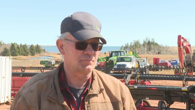 John Visser stands for an interview in a ball cap and sunglasses. Behind him is a farm yard filled with farm equipment. 