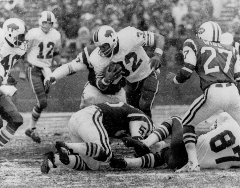 With football under one arm, a Black player runs against rival players on the field, in a black and white photo.