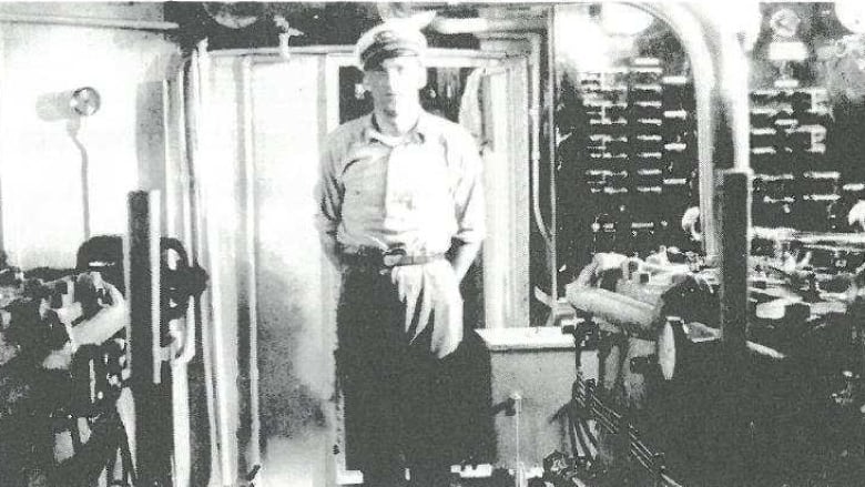 A grainy black and white photo of a man with a mariners cap in the engine room of a boat.