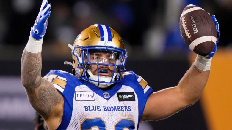 A football player in white and blue, with a gold helmet, holds up a football in celebration.