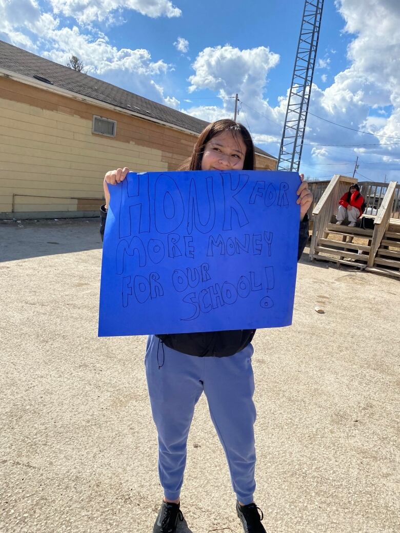 A girl holds a blue sign that reads, 'honk for more money for our school.'