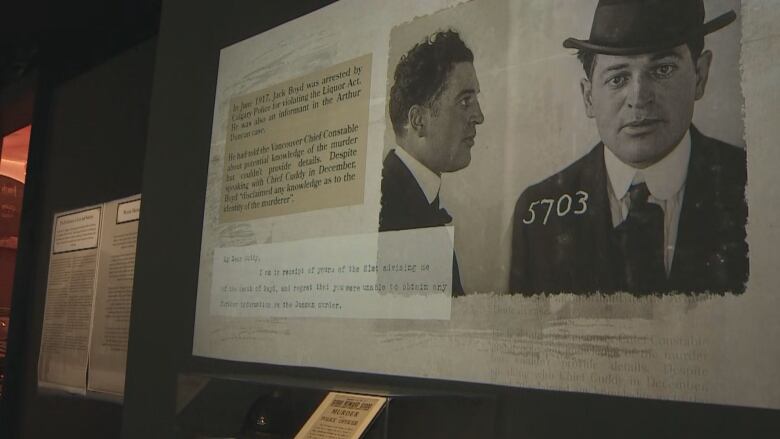 a slideshow is projected on a wall above a glass case holding artifacts. there are black and white mug shot images of a man in a hat.