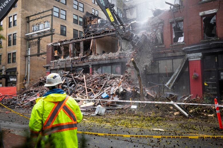 A building being demolished as a worker in reflective clothing looks on.