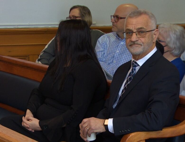 A bearded man in a business suit sits on a bench in a wide courtroom.