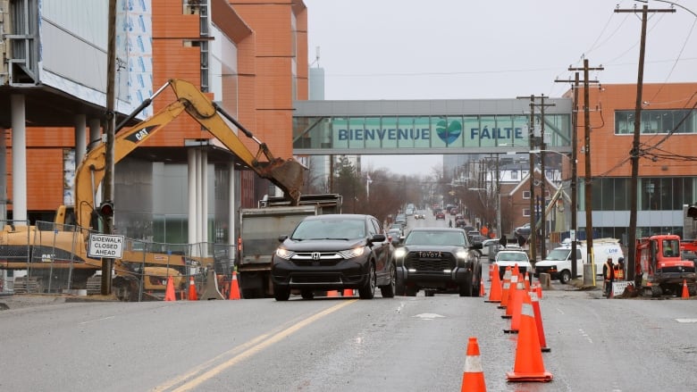 A line of cars gets squeezed into one lane by a series of fluorescent orange plastic traffic cones while construction equipment works on the street.