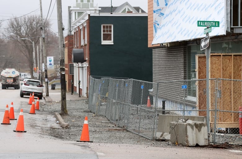 A city street is shown with a chain-link fence blocking a construction site next to a stone-covered sidewalk marked by orange plastic traffic cones.