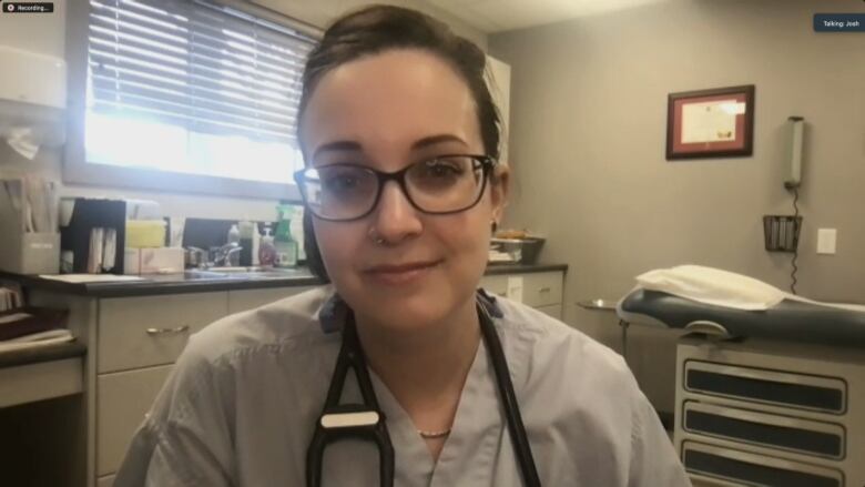 A doctor is pictured in a virtual on camera interview in an examination room wearing blue scrubs with a stethoscope around her neck.