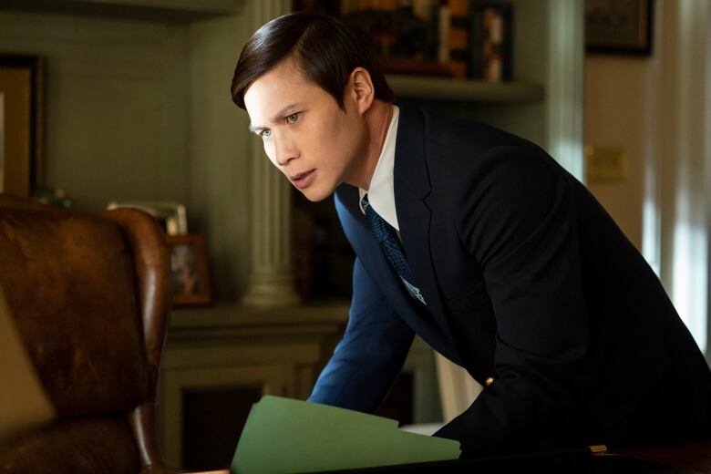 A man wearing a suit holds a file folder as he hunches over a desk. 