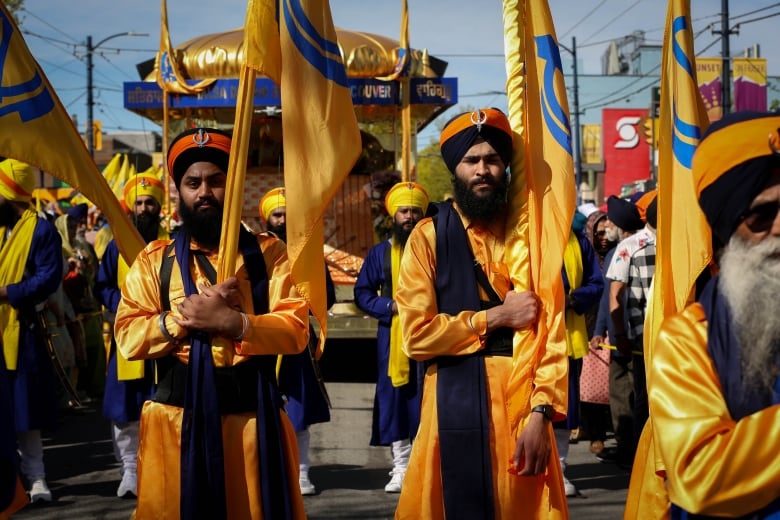 Men wearing orange regal clothes and turbans march down a street, escorting a large parade float.