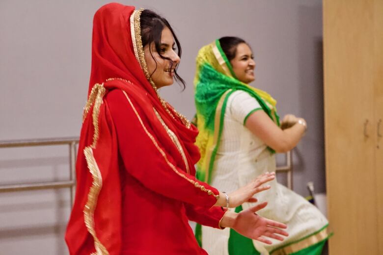 An East Indian woman in a bright red dress waves her hands in front of her as she dances, with another female dancer seen in the background. 