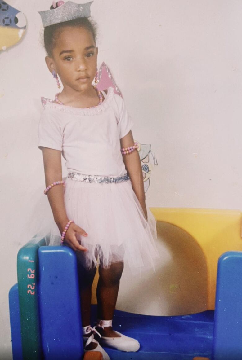 A young girl wearing a pink ballet tutu, silver crown and ballet flats poses on a blue and yellow play structure. 