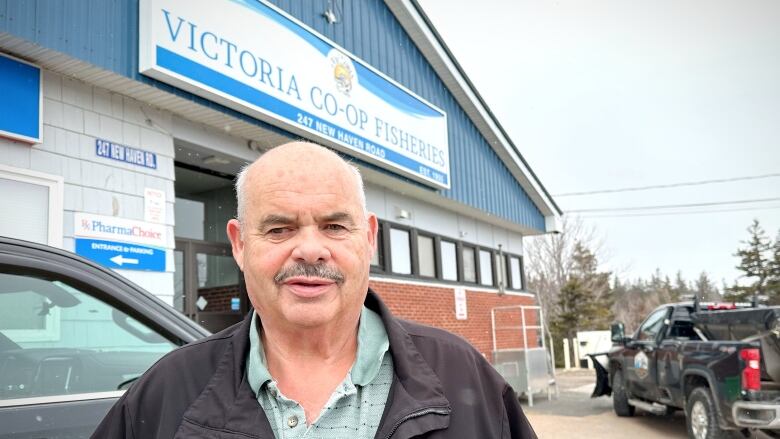 A balding man with white hair, dark grey moustache wearing a green shirt and a black jacket stands in front of a fisheries building.