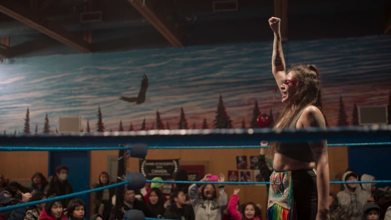 A woman with red face paint, in beadwork-style tights and a sports bra stands in a wrestling ring, fist raised.