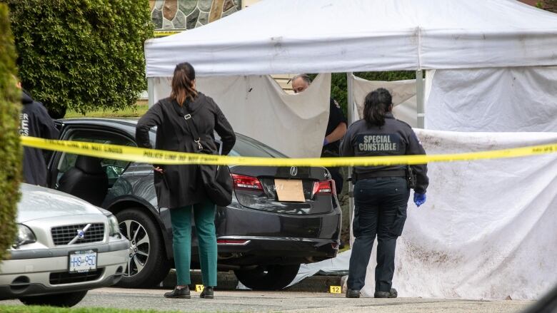 Investigators stand in front of a white tent with police tape cordoning off a black car.