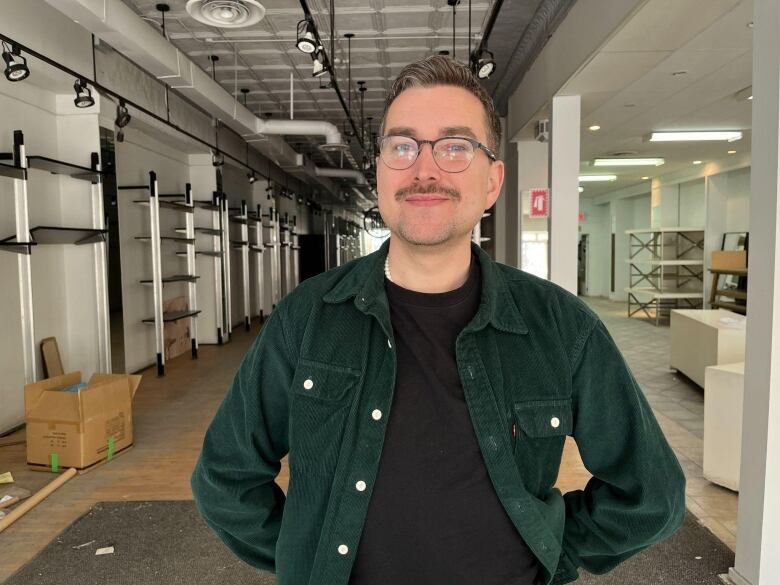 White man wearing a green jacket, black tee shirt and round glasses standing in a hallway with a faint smile.