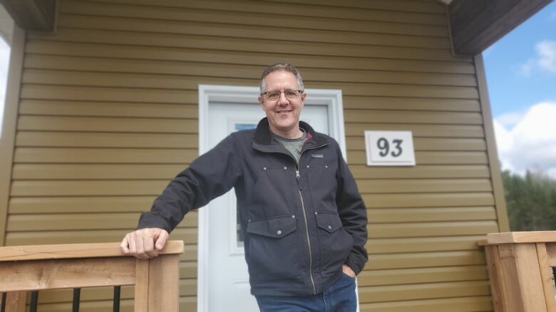 A man with salt and pepper hair wearing glasses and a black jacket smiles at the camera while leaning on the deck of a tiny home with the civic number 93 over his shoulder.