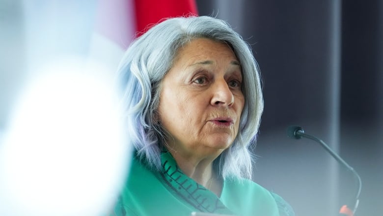 A woman in a green tunic speaks into a microphone. A glare is seen over her right shoulder.