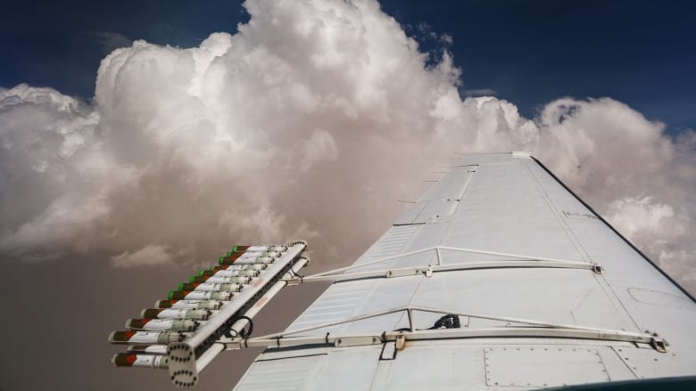 The wing of a plane with a rack of flares attached to it is seen with clouds in the background.