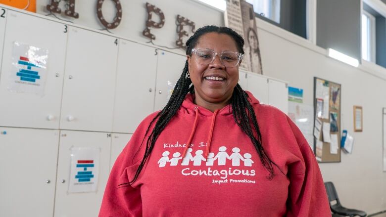 A person with long dark hair, wearing a pink hoodie, stands inside a room and smiles.