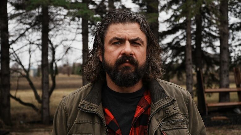 A man in a red and black plaid shirt and green jacket is picture in front of a row of trees.