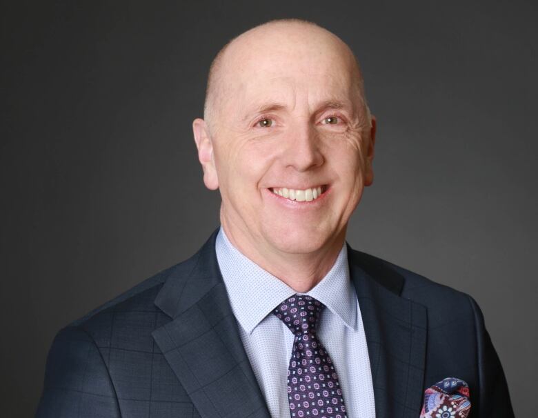 A smiling bald man wearing a suit and standing in front of a black background