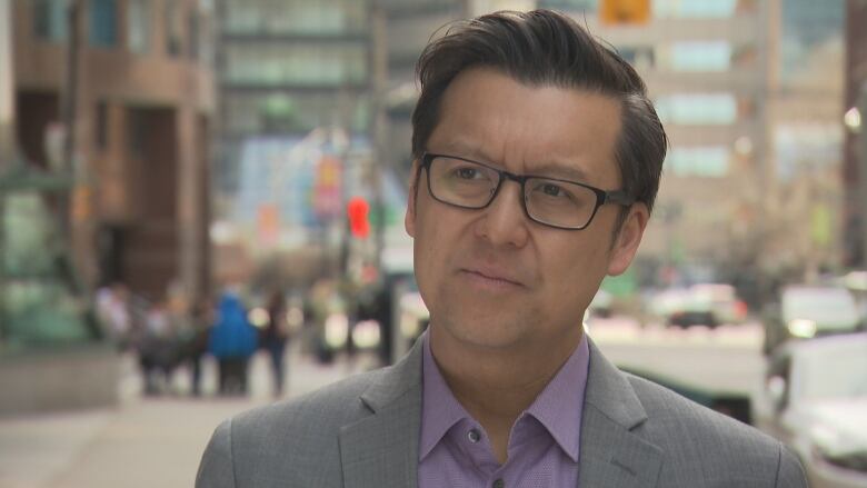 Close up on Douglas Kwan, wearing a suit and glasses, with a blurred busy downtown street corner in the background. 