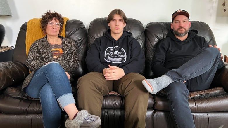 Three people sit on a brown leather couch wearing neutral facial expressions. On the left is a cross-legged woman with short brown curly hair and glasses. She is wearing jeans, a dark cardigan sweater over a grey and orange t shirt, and grey beaded moccasin style slippers. In the centre is a young man with short brown straight hair sitting straight with hands folded in his lap, brown pants, and a black hoodie sweatshirt. On the left sits an older man with one leg crossed at the knee. He has a black and grey beard, dark baseball style hat, grey jeans, and grey wool socks. 