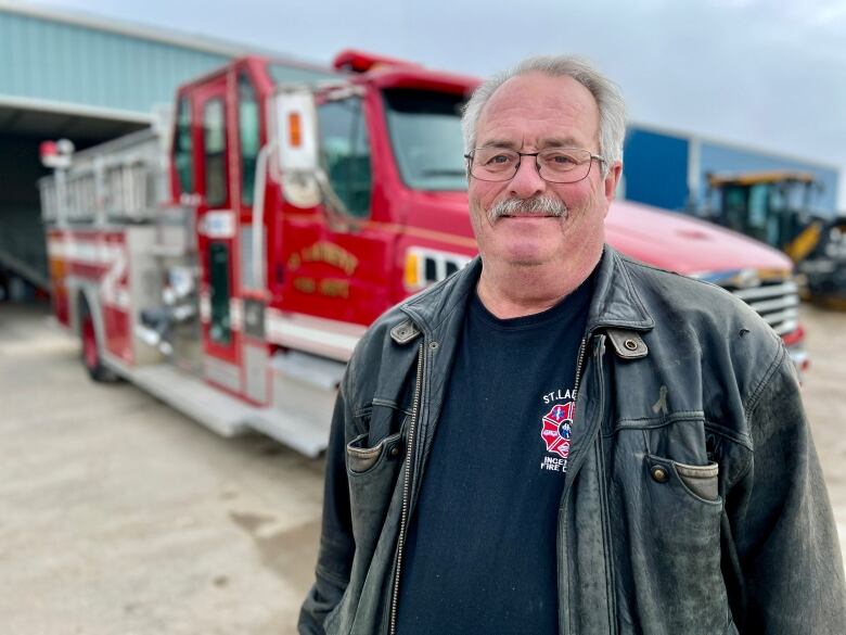 A man with grey hair and glasses stands in front of a fire engine.