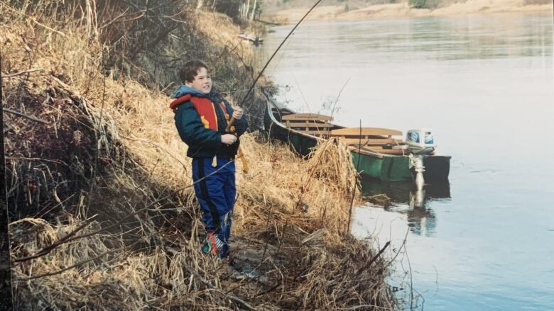 A young boy wearing life jacket, standing on river bank and pulling up on a fishing rod