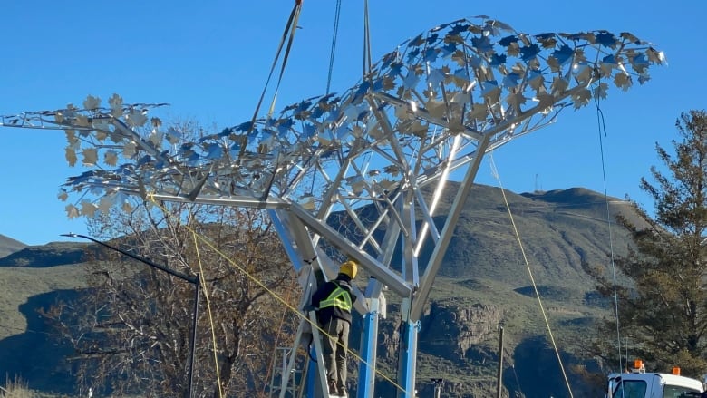 A stainless steel outline of a plane is mounted on steel pillars by construction workers. There is a mountain range in the background.