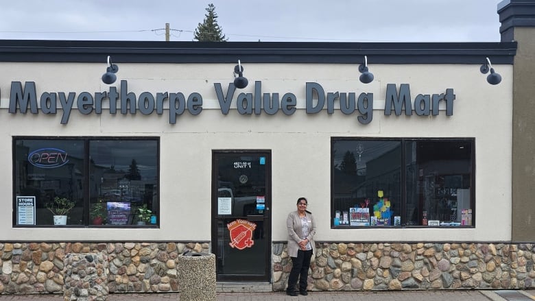 A woman folds her hands standing in front of a storefront saying Mayerthorpe Value Drug Mart. 