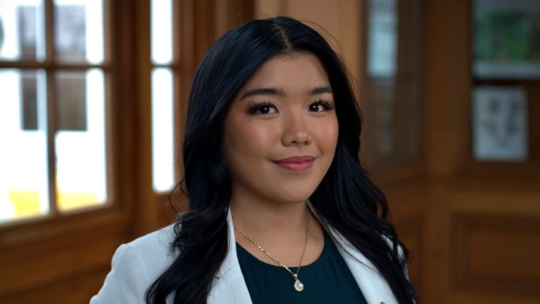 A young woman with red lipstick who is wearing white pants and a white coat looks at the camera.