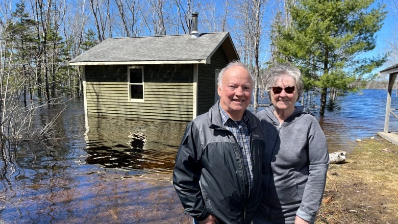 Two people stand in front of a body of water that's surrounded a small building in the background.