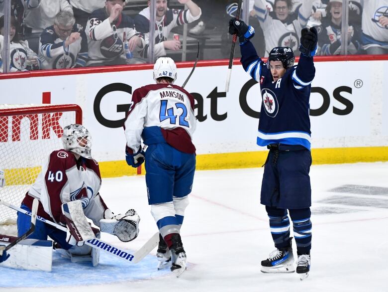 A hockey player raises his arms as a goaltender looks on from his net.