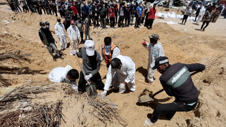 People in white hazmat suits use shovels to dig through brown, sandy dirt.