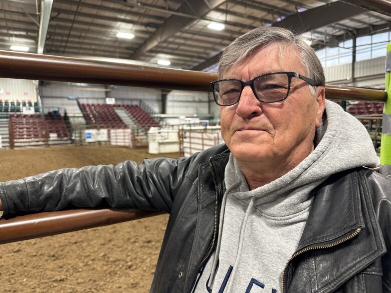 A man wearing a leather jacket stands with his arm on the rail outside a dirt arena. 