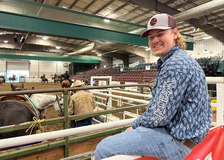 A man wearing a baseball cap sits on a rail fence on the edge of a rodeo arena.