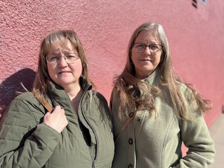 Two women stand next to each other, both wearing green coats and glasses.