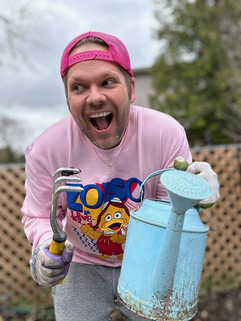 A man with a watering can an a rake smiles while looking off to the side with a fence in the background